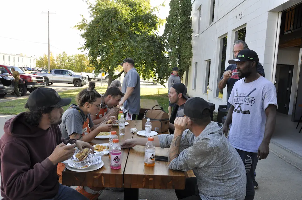 5 people sitting at a picnic table eating burgers and laughing with each other.