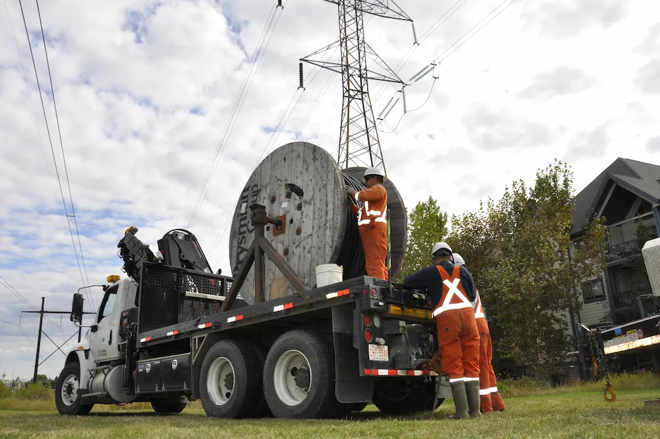 Employees unloading a giant ream of cable from the back of a truck with power lines in the background.